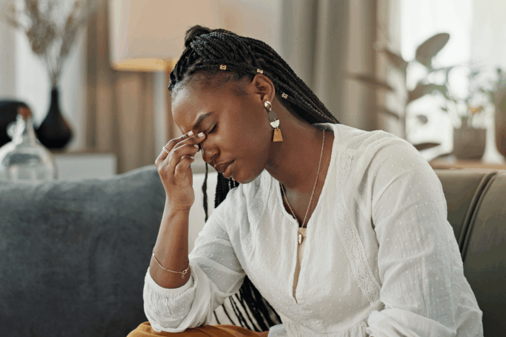 A woman sat on the sofa looking stressed. She is holding the bridge of her nose.