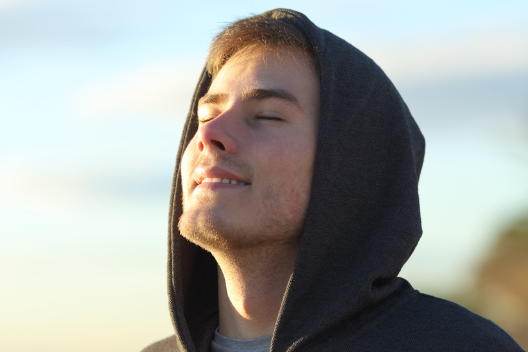 A young person is wearing a hoody. He has his eyes closed and is basking in the sunlight